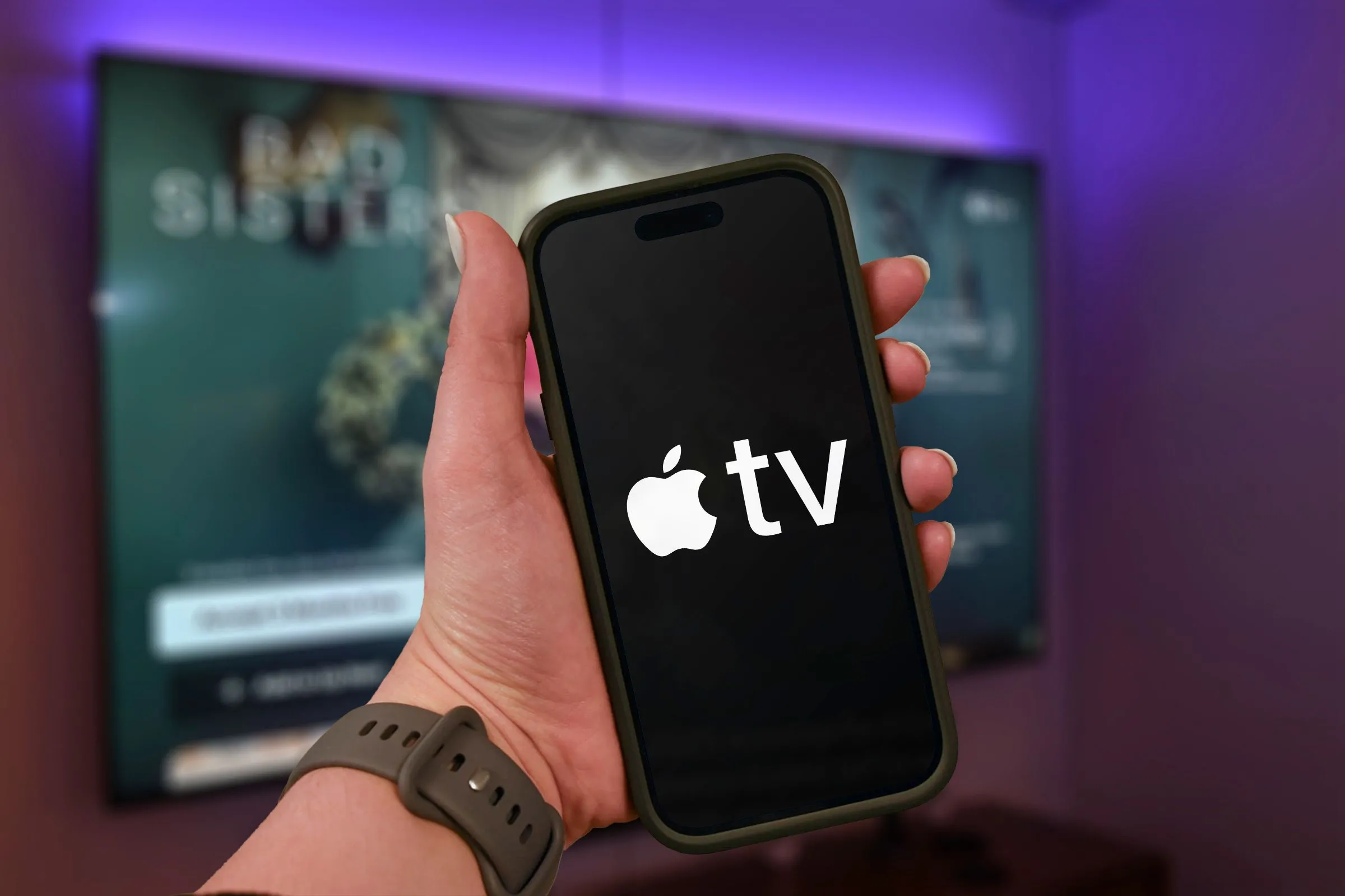Người dùng đang cầm iPhone hiển thị logo Apple TV+ với TV mờ phía sau