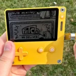 Người dùng đang chơi game Pick Pack Pup trên máy chơi game cầm tay Playdate, nổi bật tay quay độc đáo bên cạnh.