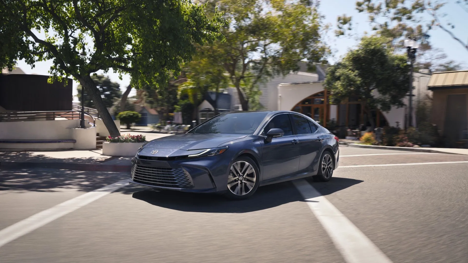 Ô tô Toyota Camry 2026 màu xám bạc di chuyển trên đường phố, thể hiện thiết kế "đầu búa" và đèn LED hiện đại.